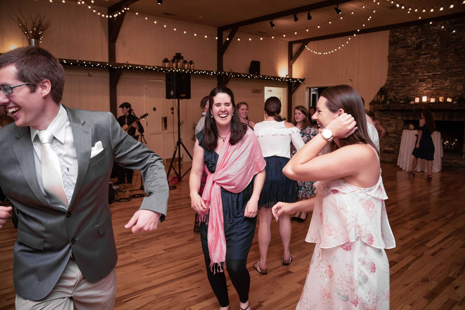 Guests dancing joyfully at a wedding reception in a rustic venue with string lights overhead.