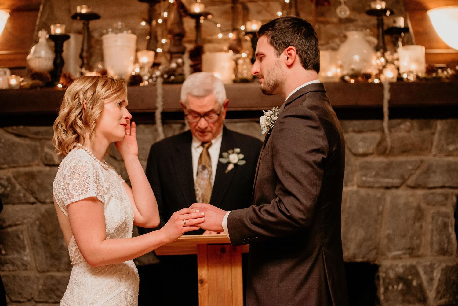 Emotional bride wiping tears during indoor wedding ceremony at rustic venue with candlelit backdrop.