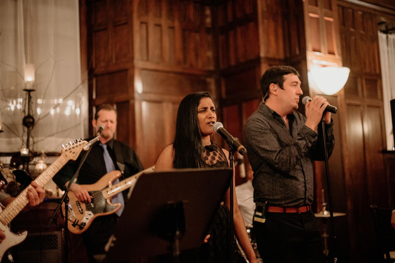 Live band performing at elegant wood-paneled venue, featuring guitarist and two vocalists singing into microphones.