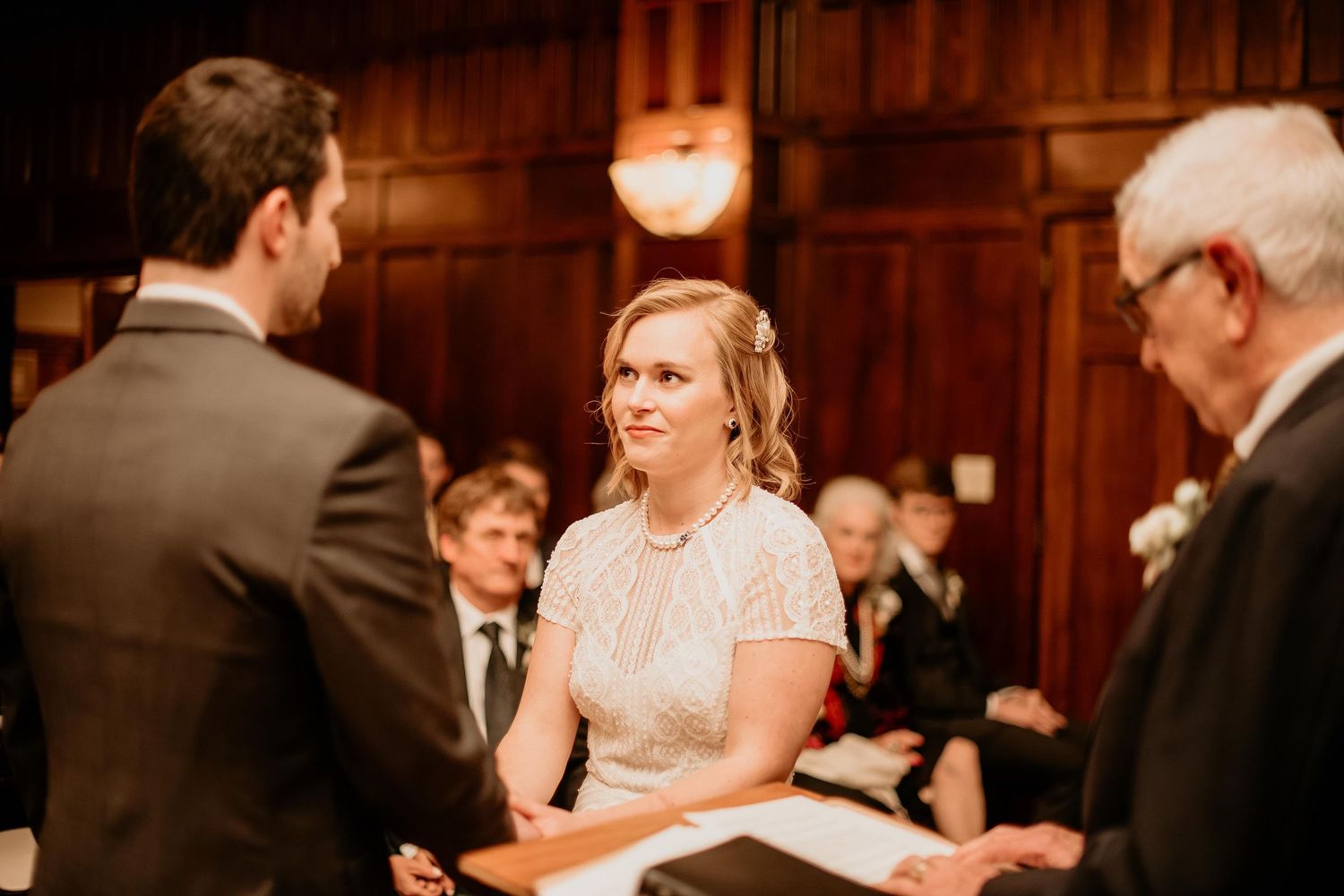 Bride smiling at groom during indoor wedding ceremony, officiant visible at right, guests seated in background.