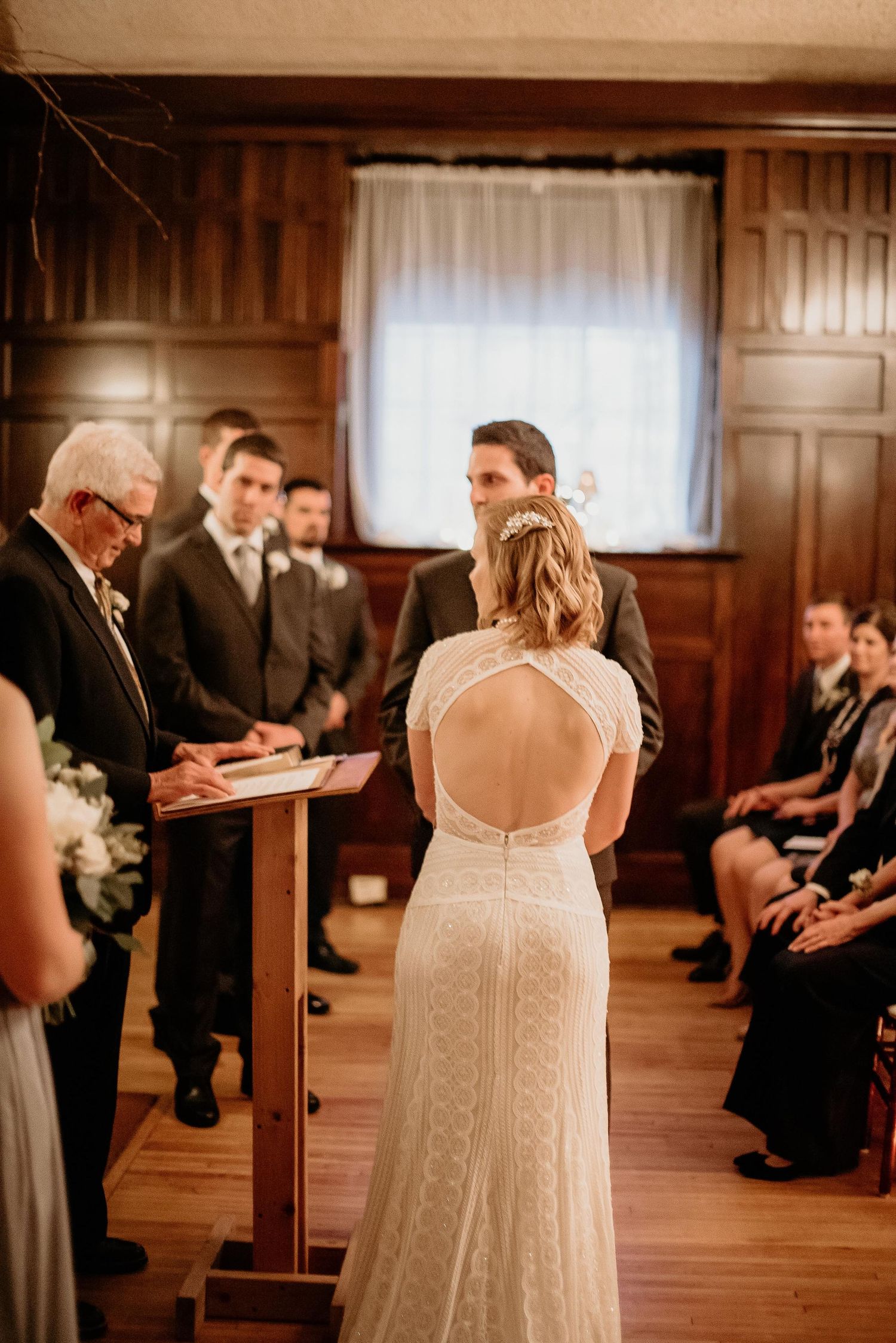 Bride in lace open-back gown faces groom during indoor wedding ceremony with wood-paneled walls.