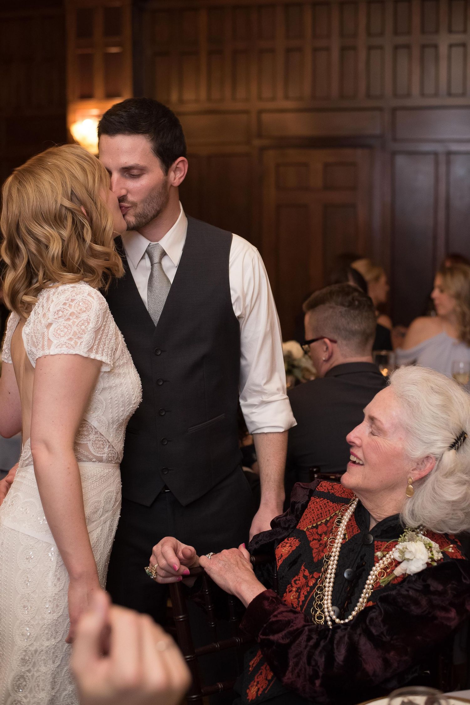 Bride and groom share a kiss at wedding reception while elderly woman in pearls smiles warmly nearby.