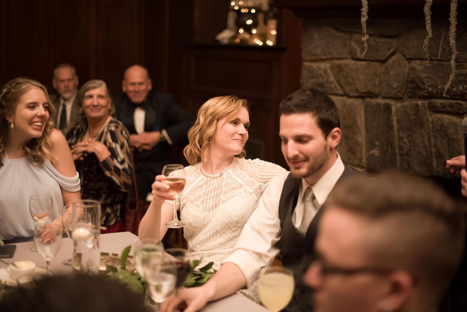 Bride and groom smiling at wedding reception dinner table, guests celebrating in elegant stone fireplace venue.