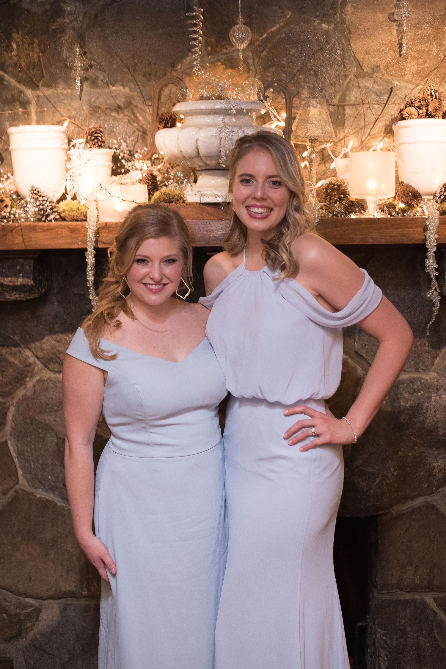Two women in matching light blue gowns pose together in front of a stone fireplace with elegant holiday decor.