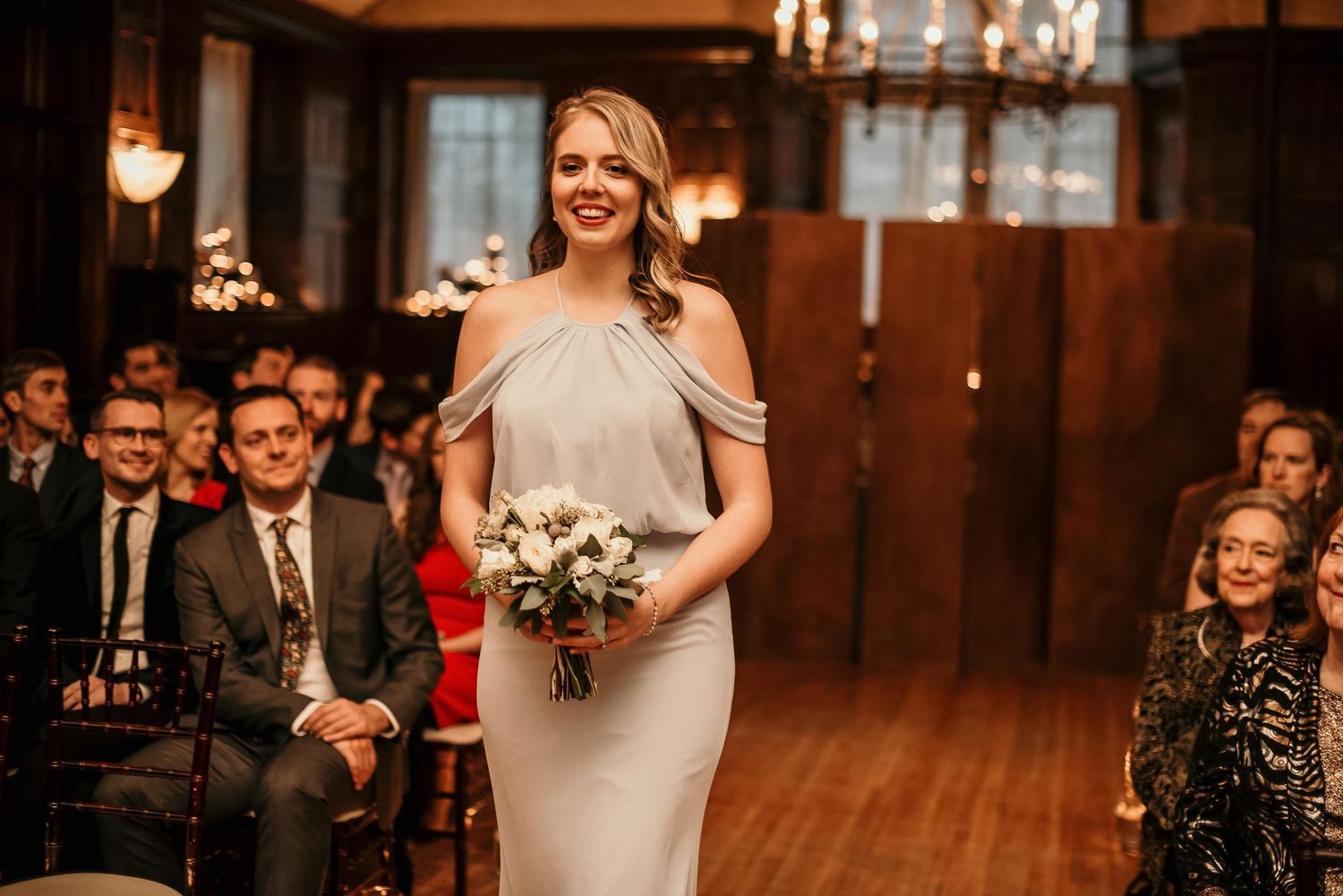 Bridesmaid in silver gown walks down aisle holding white bouquet at elegant indoor wedding ceremony.