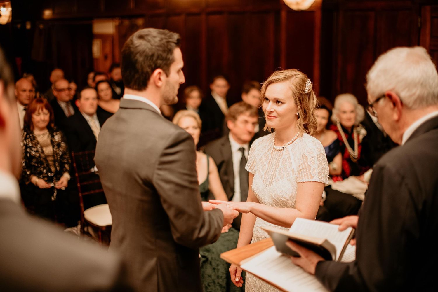 Bride and groom exchange vows during indoor wedding ceremony with guests seated in background.