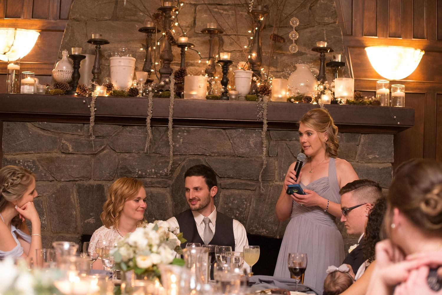 Woman giving a speech at an elegant wedding reception near a stone fireplace with candles and floral arrangements.