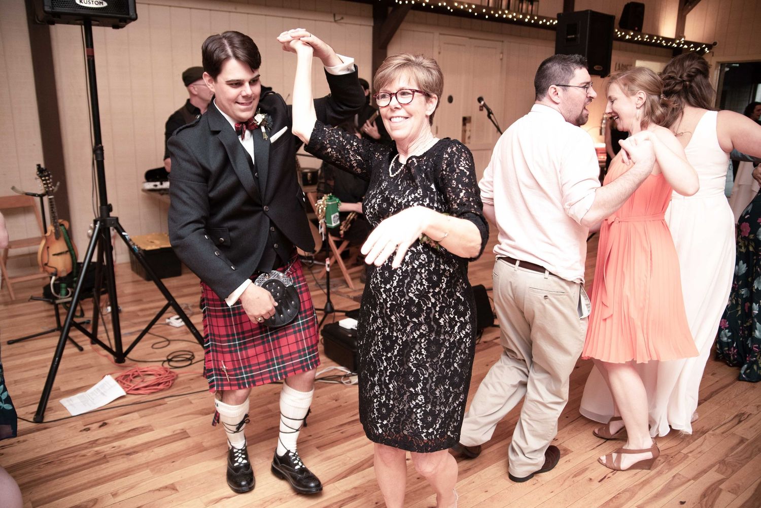 Man in Scottish kilt dancing with woman in black lace dress at lively wedding reception with couples dancing in background.