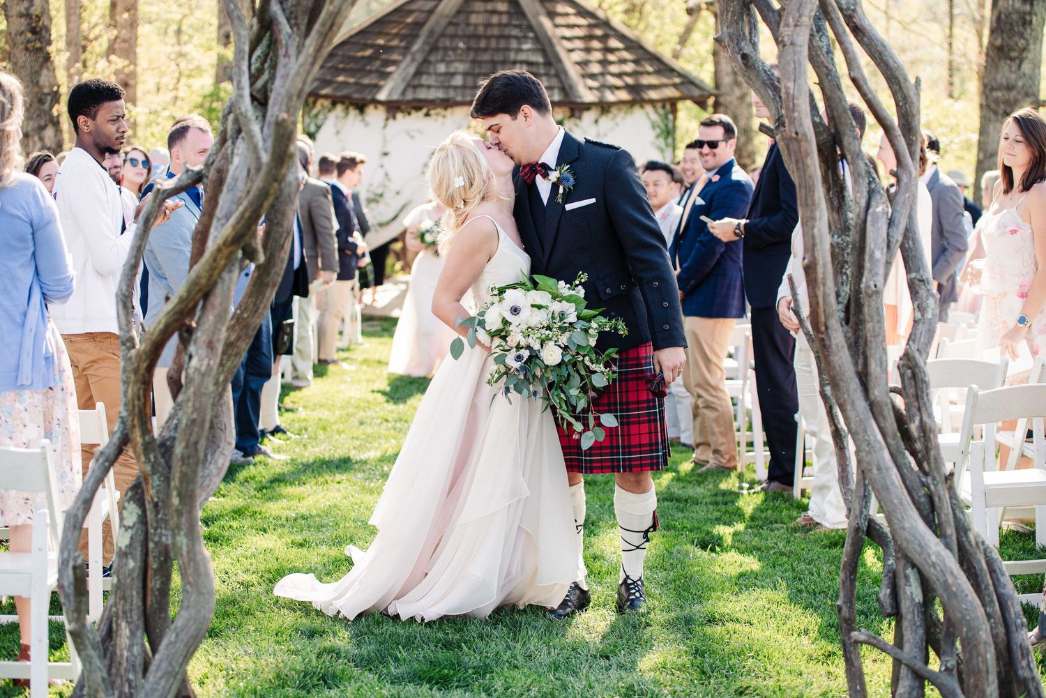 A bride and groom share a kiss under a rustic arch at an outdoor wedding ceremony with guests.