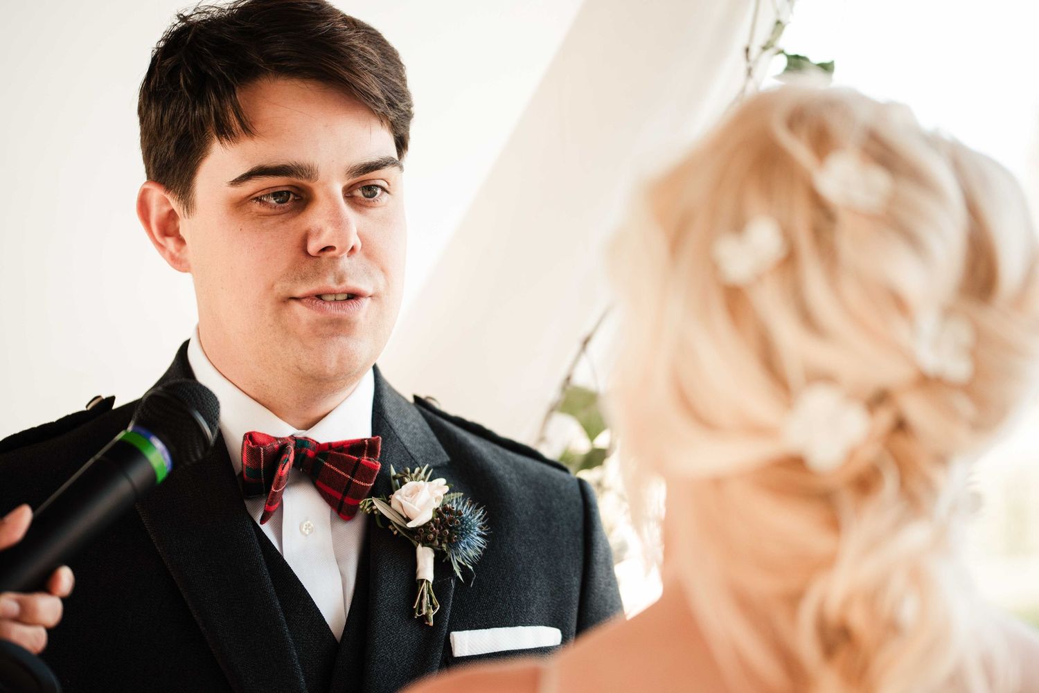 Groom in black suit with plaid bow tie and floral boutonniere recites vows to blonde bride.