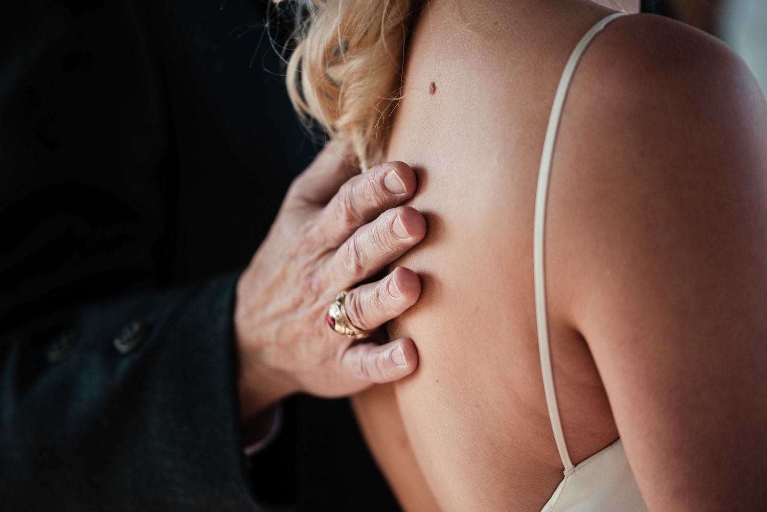 Close-up of a hand with a gold ring resting on a woman's bare shoulder in a wedding setting.