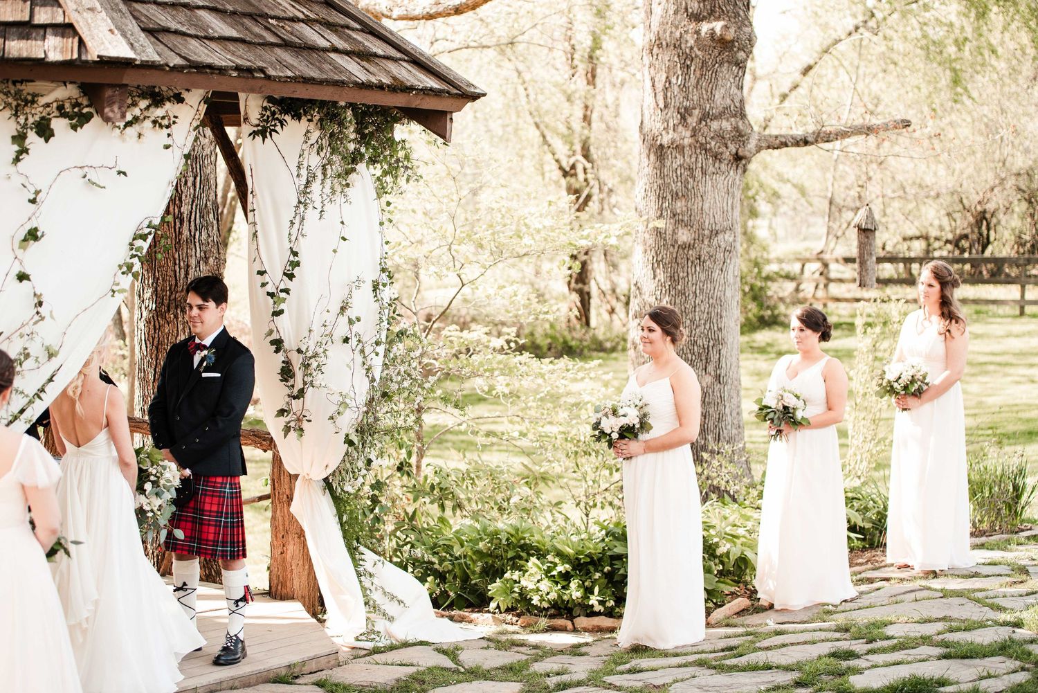 Outdoor wedding ceremony with bridesmaids in white gowns and groom in red tartan kilt standing under floral arch.