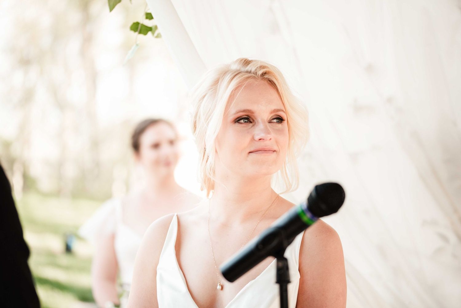 Emotional bride listening to vows at outdoor wedding ceremony with bridesmaid smiling in background.