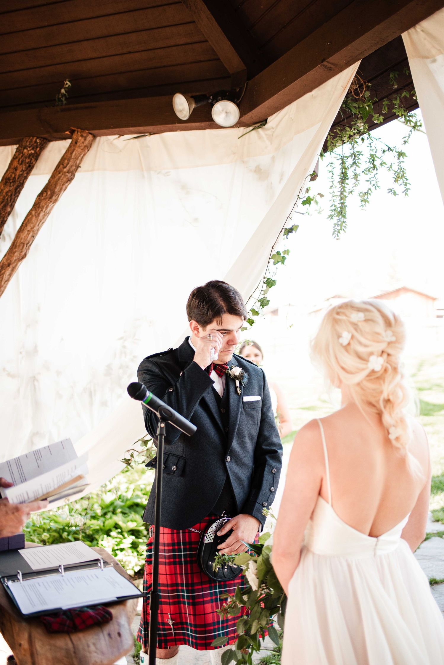 Emotional groom in tartan kilt wiping tears during outdoor wedding ceremony while bride stands before him.