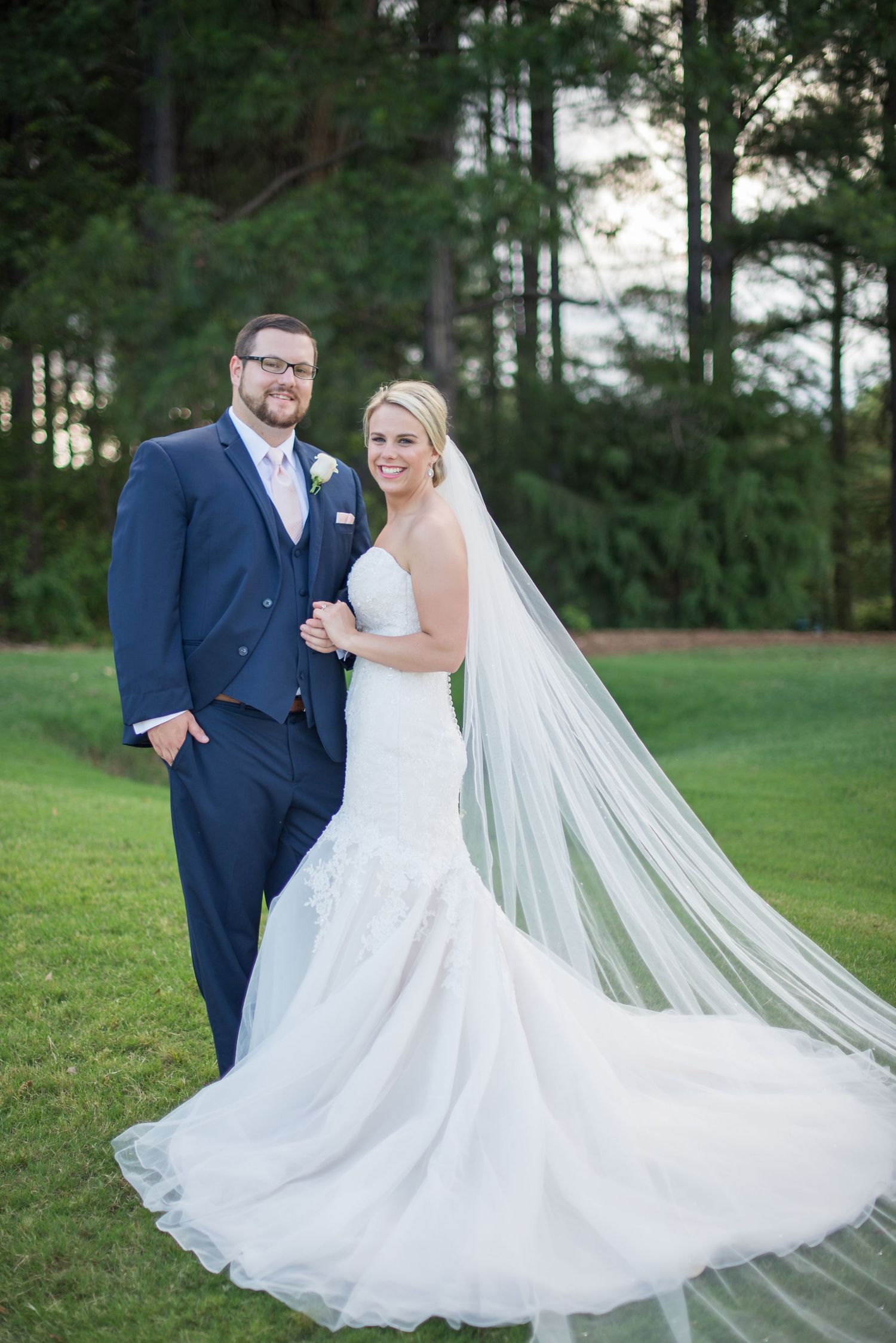 Bride in white mermaid gown with cathedral veil poses with groom in navy blue suit on green lawn.