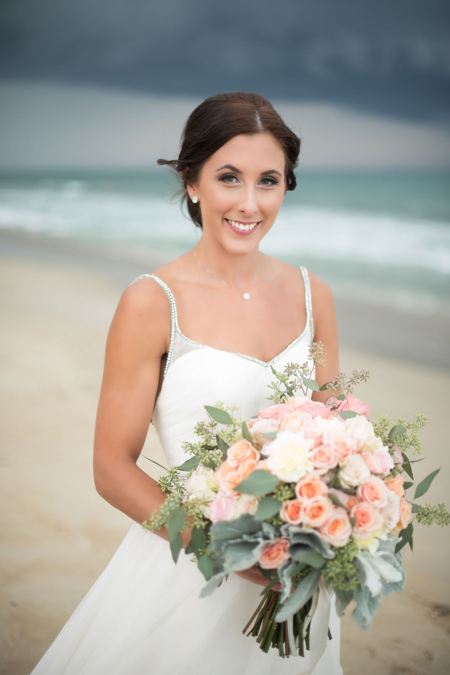 Smiling bride in white gown holding pink and peach bouquet on a beach with stormy skies.