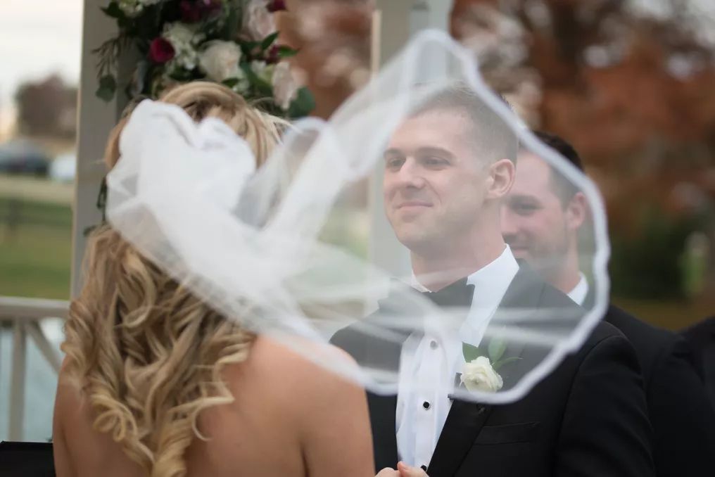 Groom smiling at bride during outdoor wedding ceremony with veil blowing in the wind.