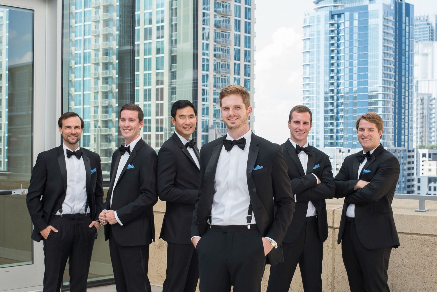 Groom and groomsmen in black tuxedos posing on a rooftop with city skyline backdrop.