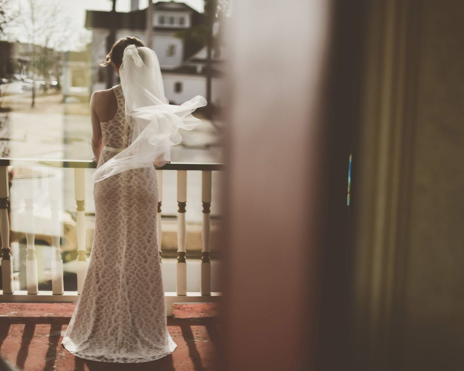 Bride in lace wedding gown stands on balcony with veil blowing in wind.