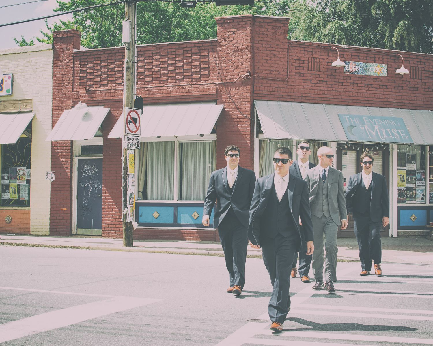 Groomsmen in dark suits and sunglasses walk confidently across a city street on wedding day.