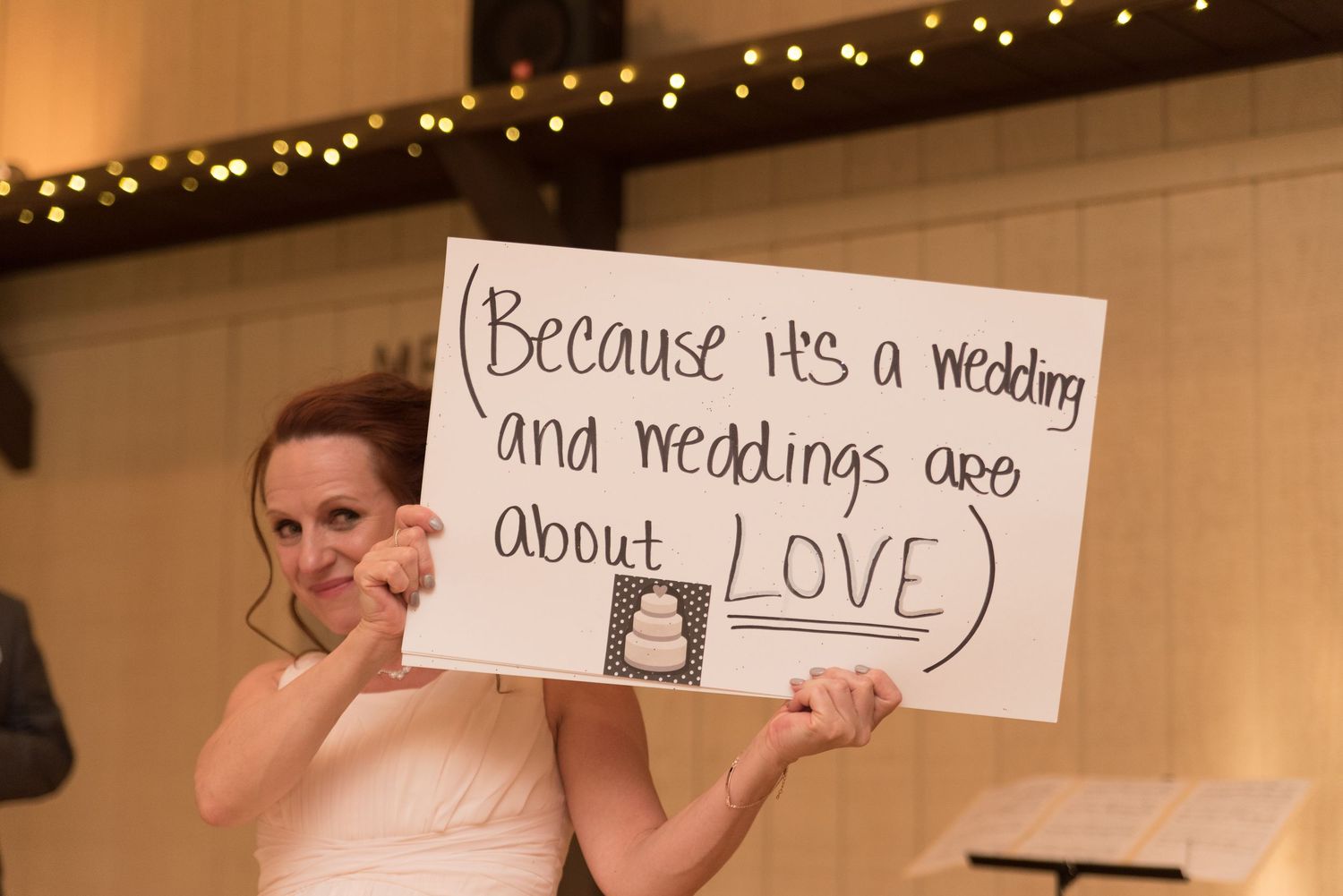 Bride holds sign reading because it's a wedding and weddings are about love at reception.