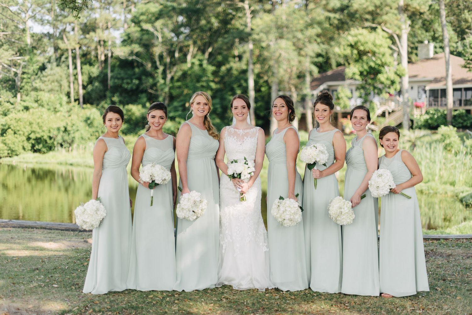 Bride with bridesmaids in mint green gowns holding white bouquets beside a serene pond outdoors.
