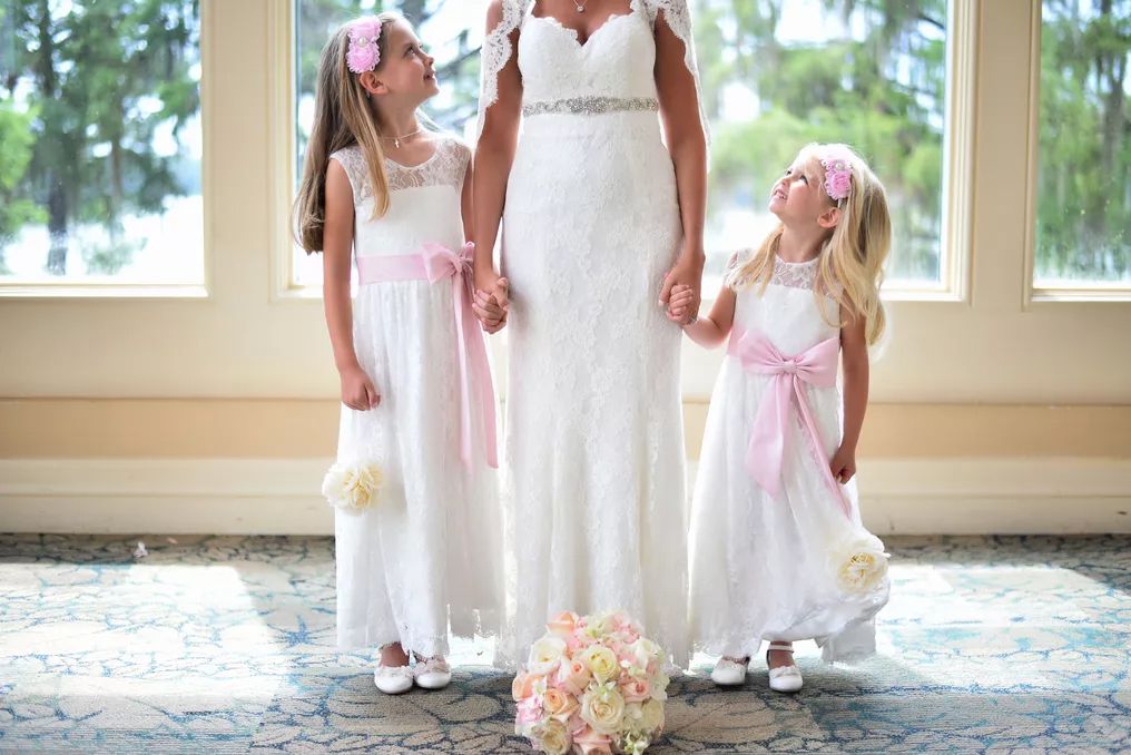 Bride in lace wedding gown poses with two flower girls in white dresses with pink sashes holding bouquet.