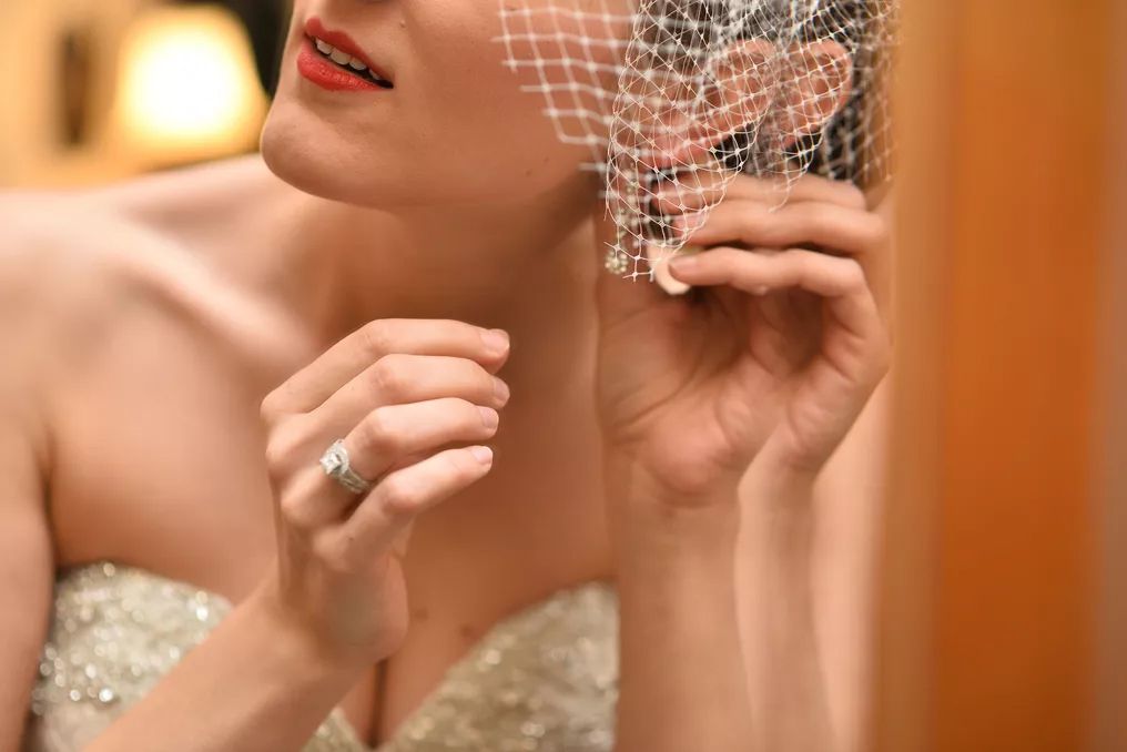 Bride adjusts birdcage veil wearing sparkling engagement ring in elegant bridal getting ready moment.