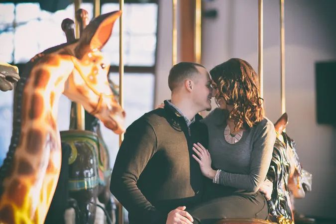 Couple sharing a romantic kiss on a carousel beside a giraffe figure at an indoor amusement venue.