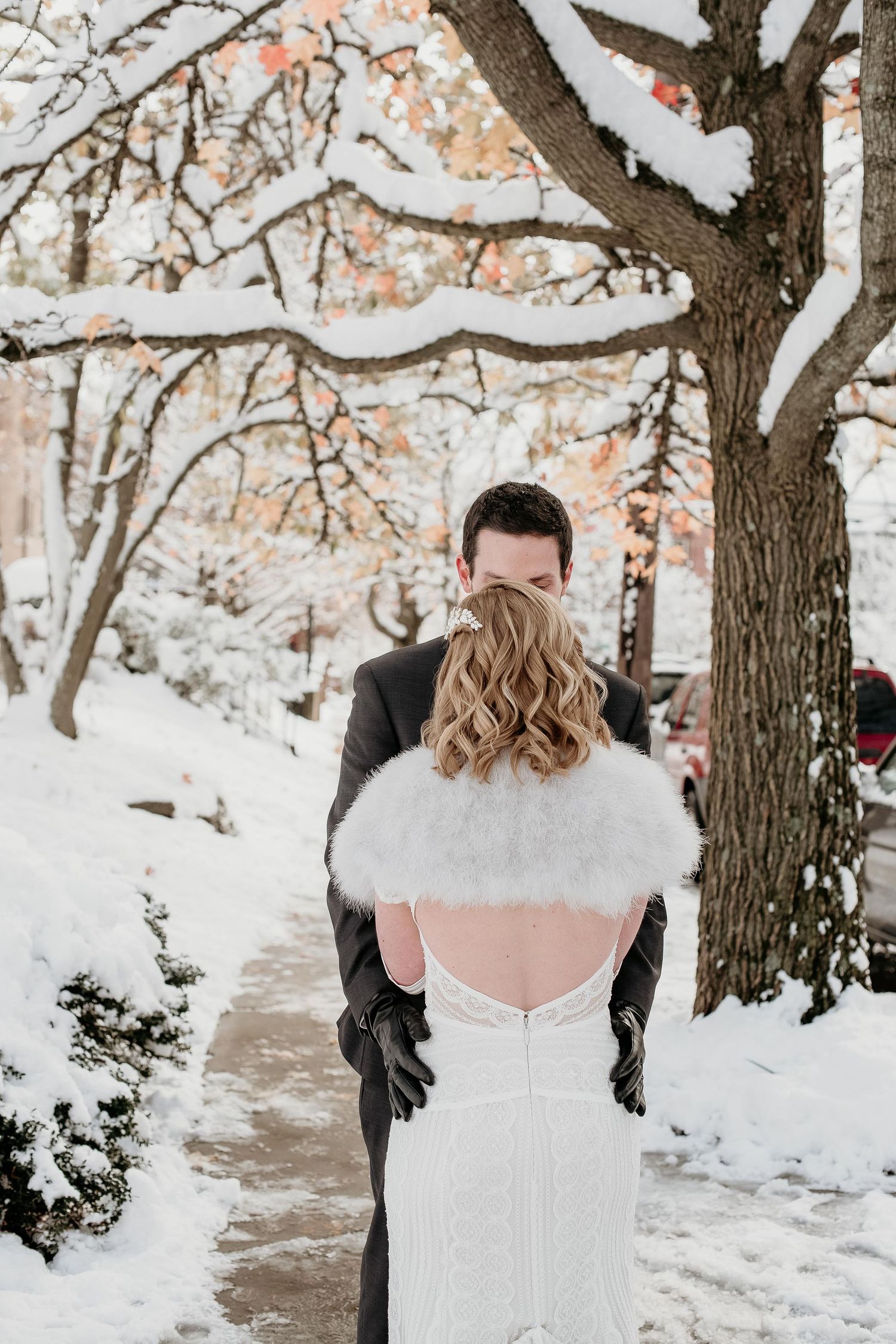 Groom kissing bride wearing white lace gown and fur shawl on snow-covered path during winter wedding.