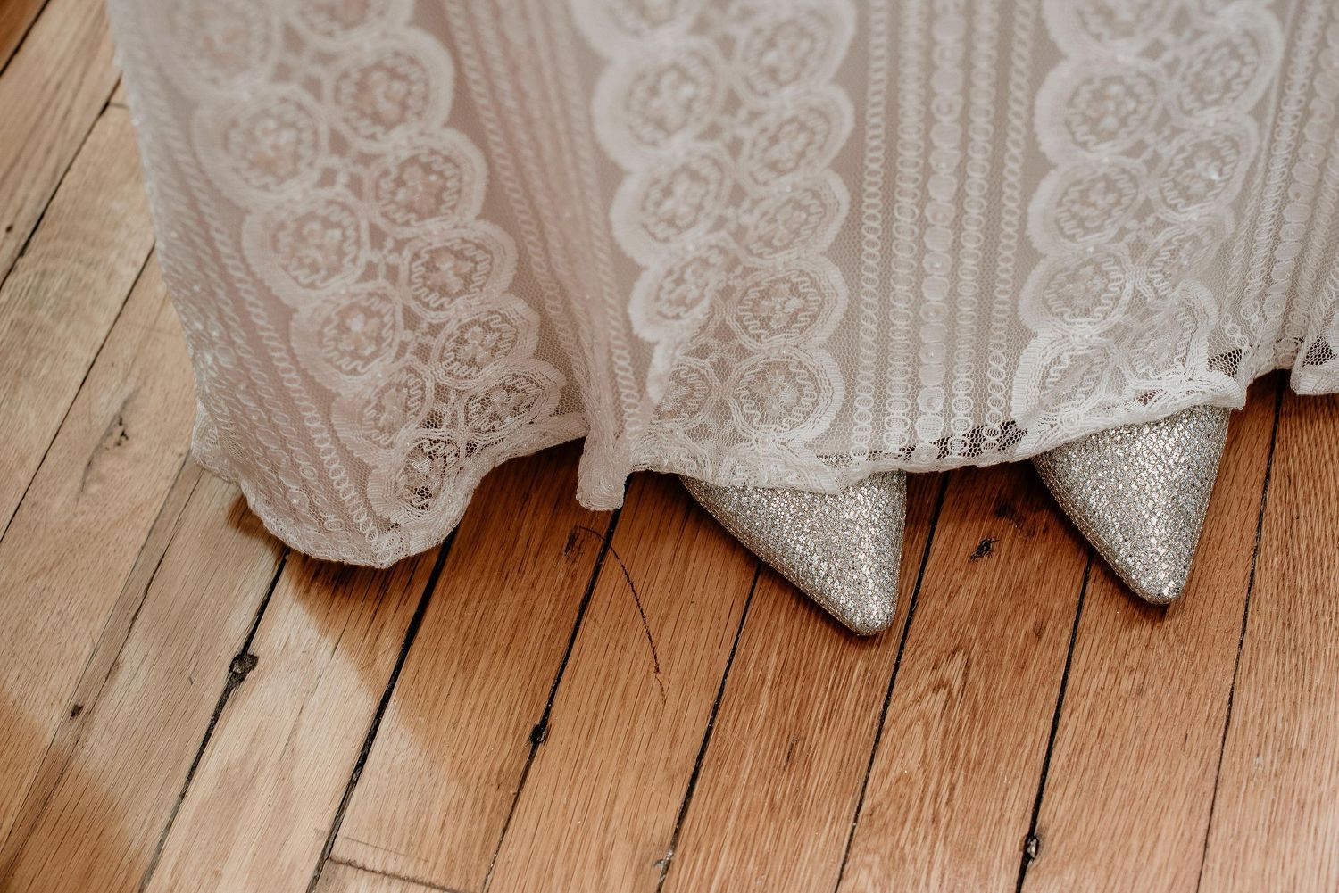 Close-up of sparkling silver bridal heels peeking beneath a lace wedding dress hem on rustic hardwood floors.