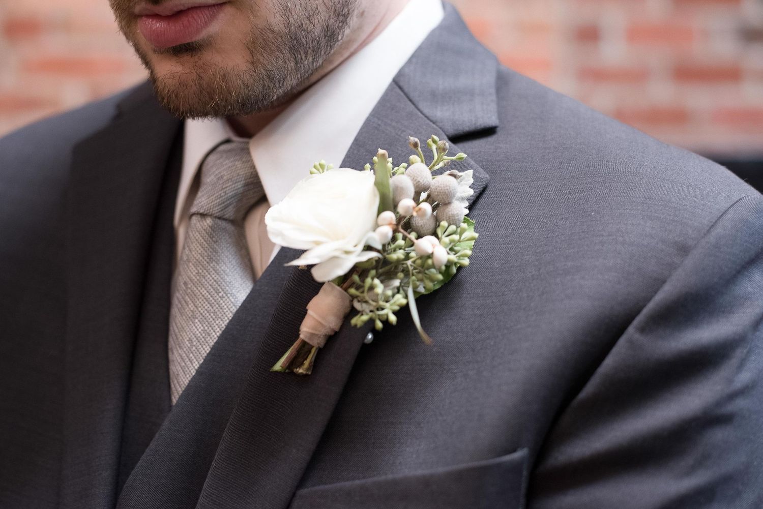 Groom wearing gray suit with white rose and greenery boutonniere pinned to lapel, paired with a tan tie.