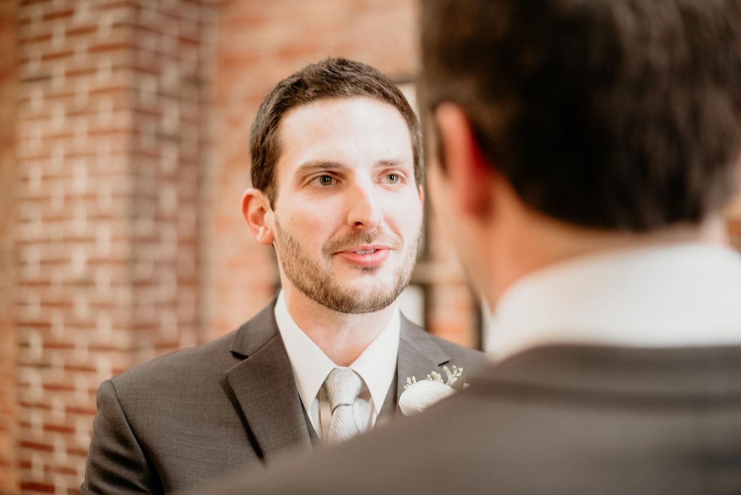 Groom in gray suit with boutonniere smiling during wedding ceremony at brick venue.