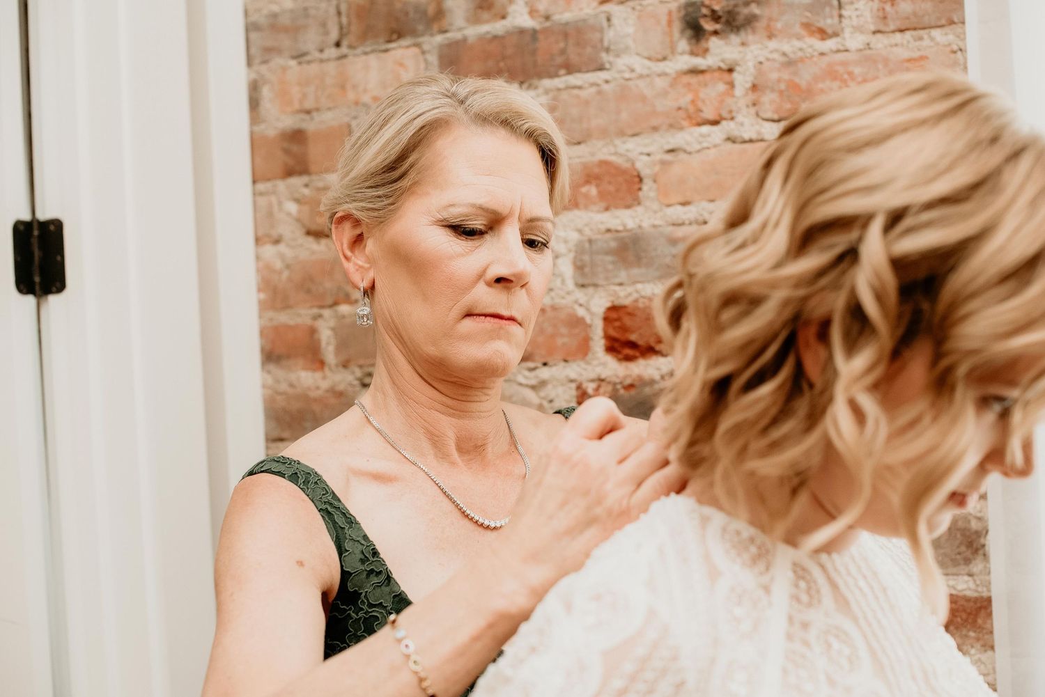Mother of the bride in green lace dress adjusting bride's hair before wedding ceremony near brick wall.