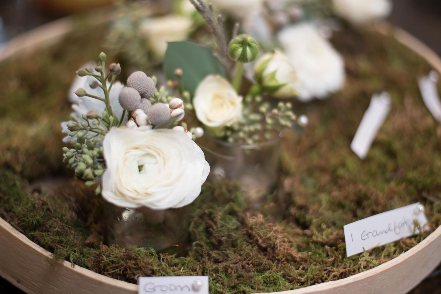 White ranunculus and berries arranged on moss in a wooden tray with name cards for a rustic wedding display.