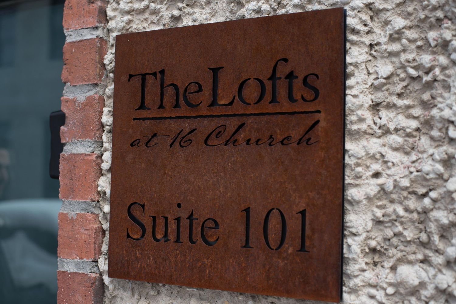 Rustic metal sign for The Lofts at 16 Church Suite 101 mounted on a brick and stone wall.