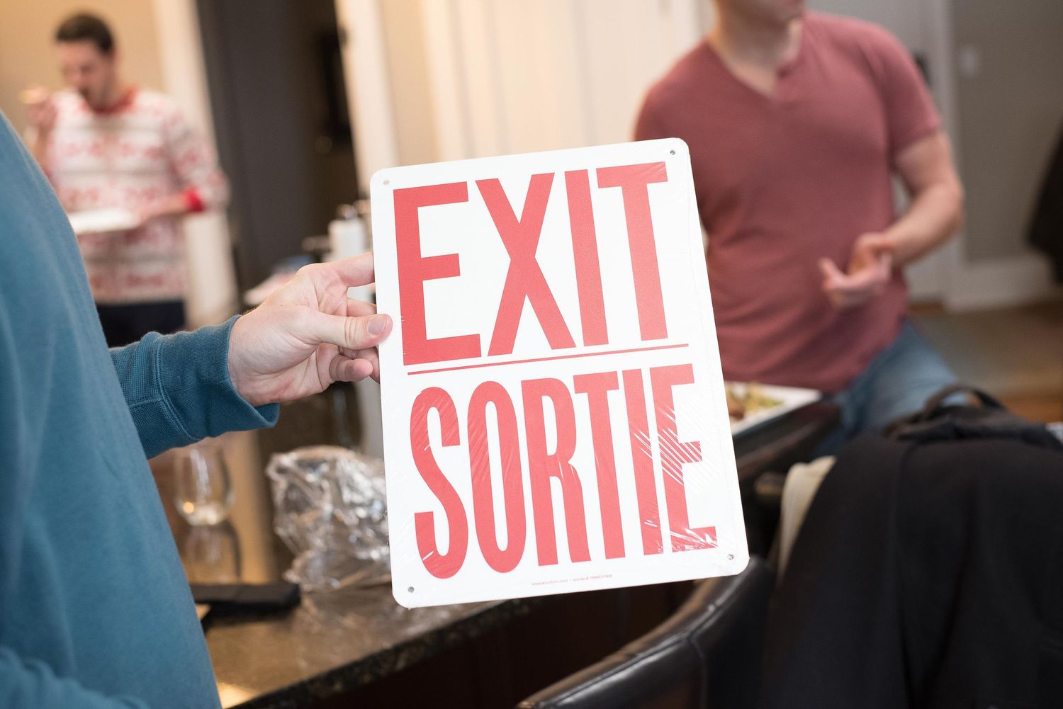Person holding a bilingual EXIT SORTIE sign in English and French inside a room with people in the background.