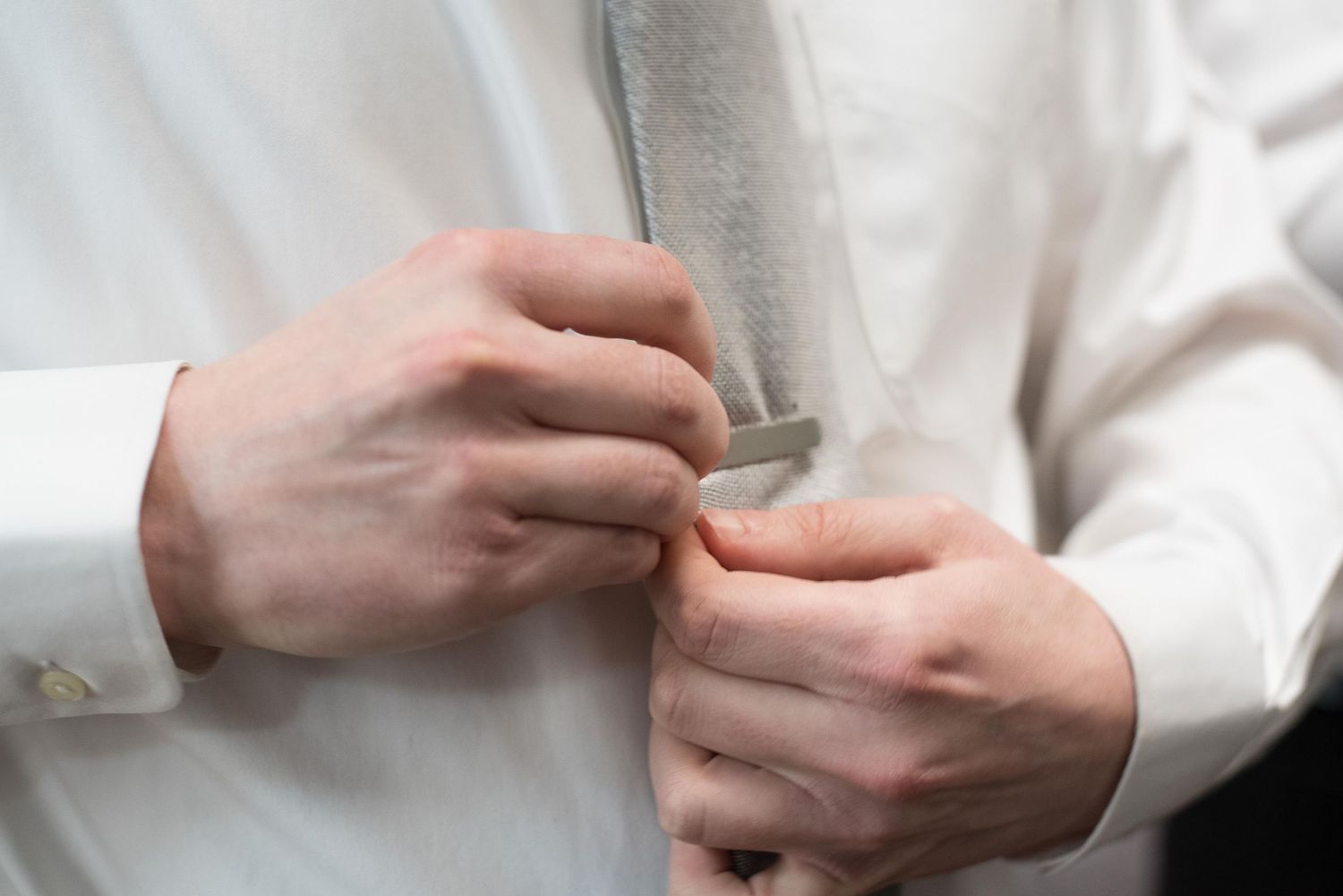 Man in white dress shirt adjusting a gray tie, close-up of hands fastening formal attire.