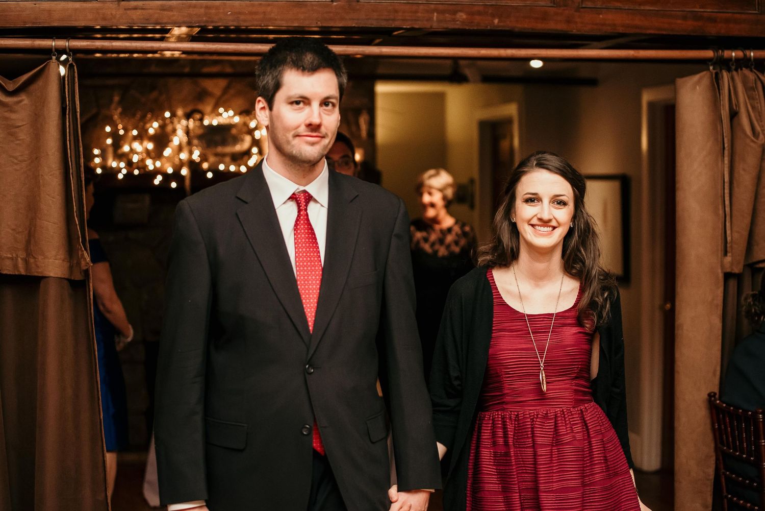 Couple in formal attire, man in black suit with red tie, woman in burgundy dress, holding hands at elegant venue.