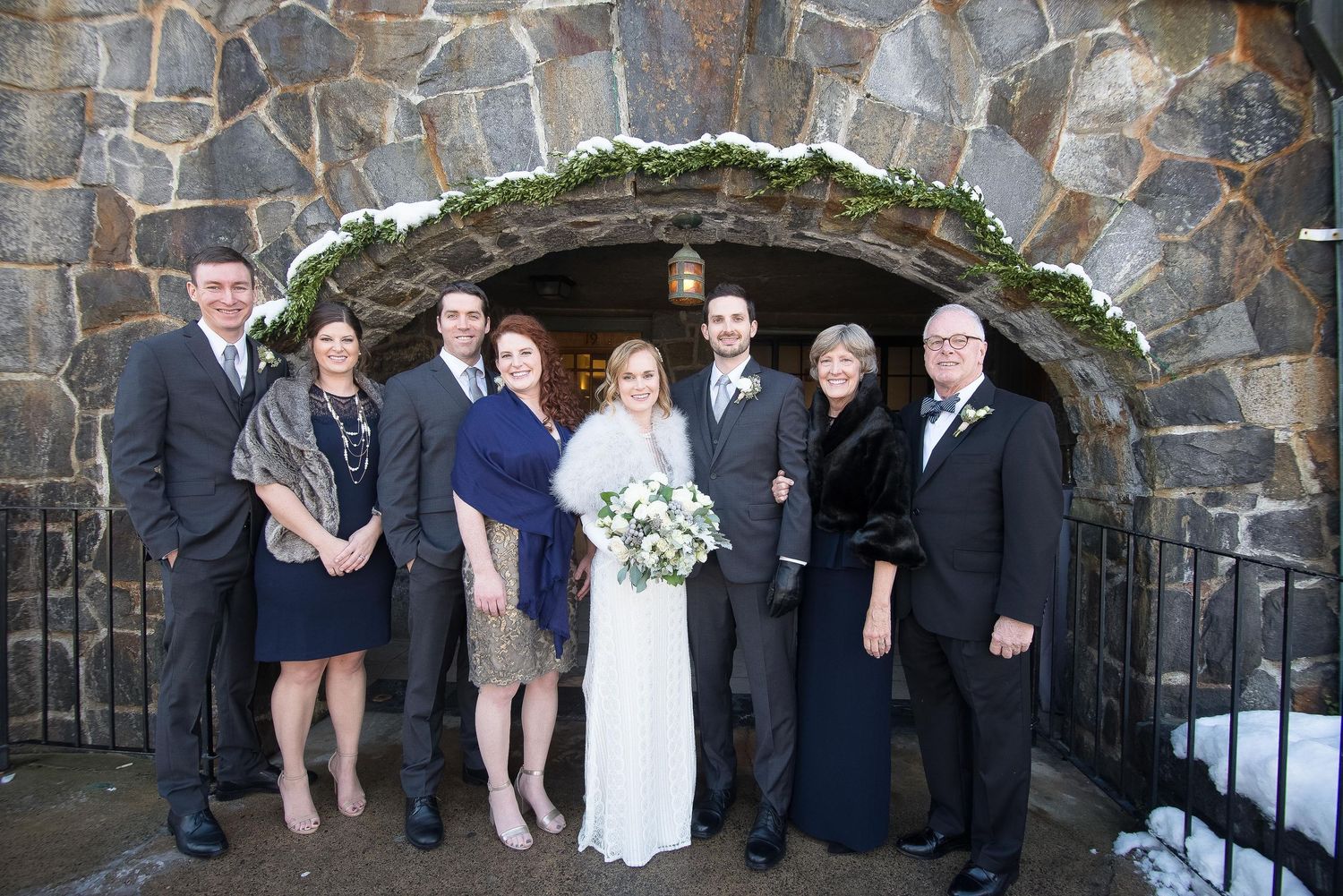 Wedding party poses in front of a stone arch entrance dusted with snow at a winter ceremony.