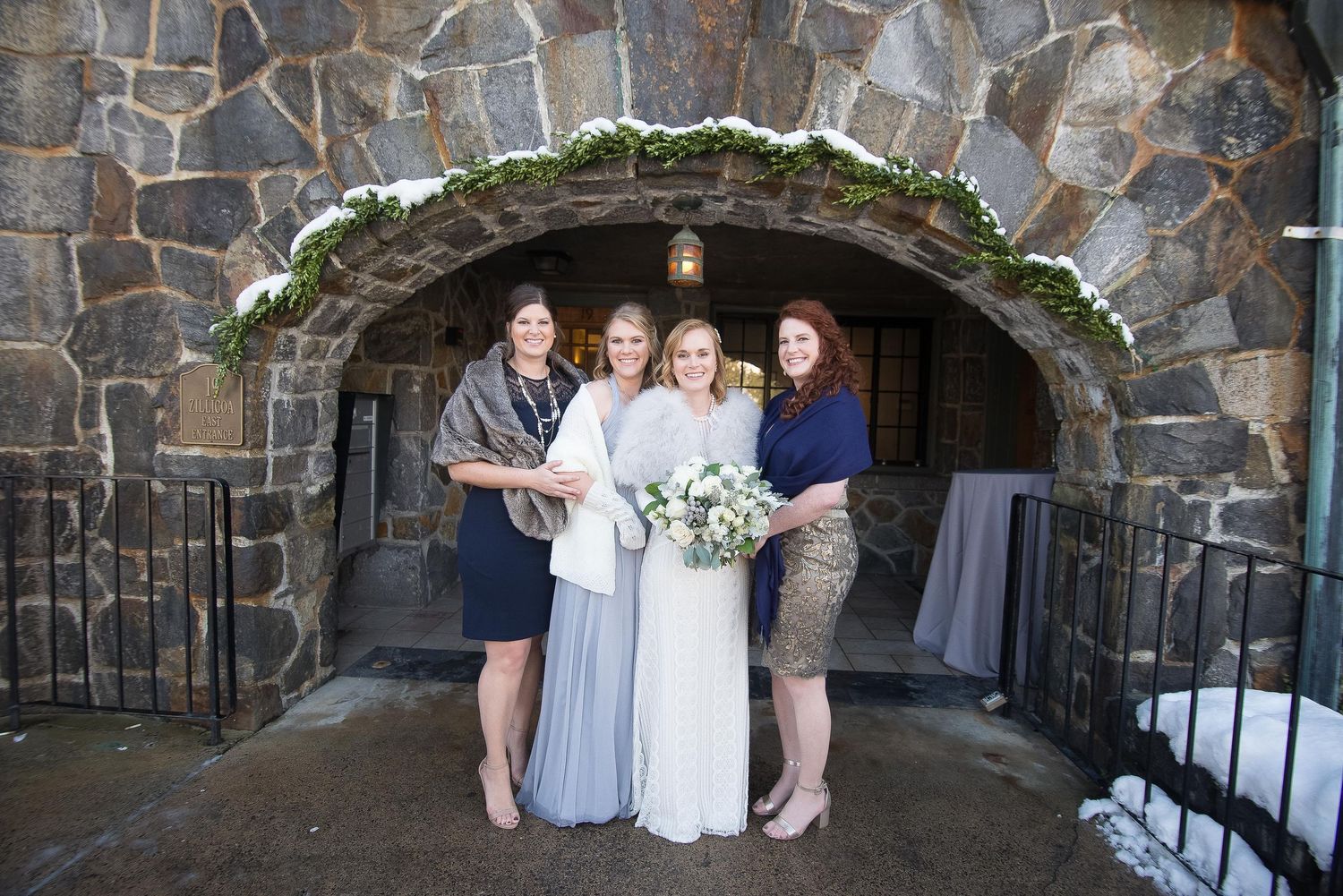 Bride with bouquet and bridesmaids in navy and gray dresses pose under stone arch at winter wedding venue.