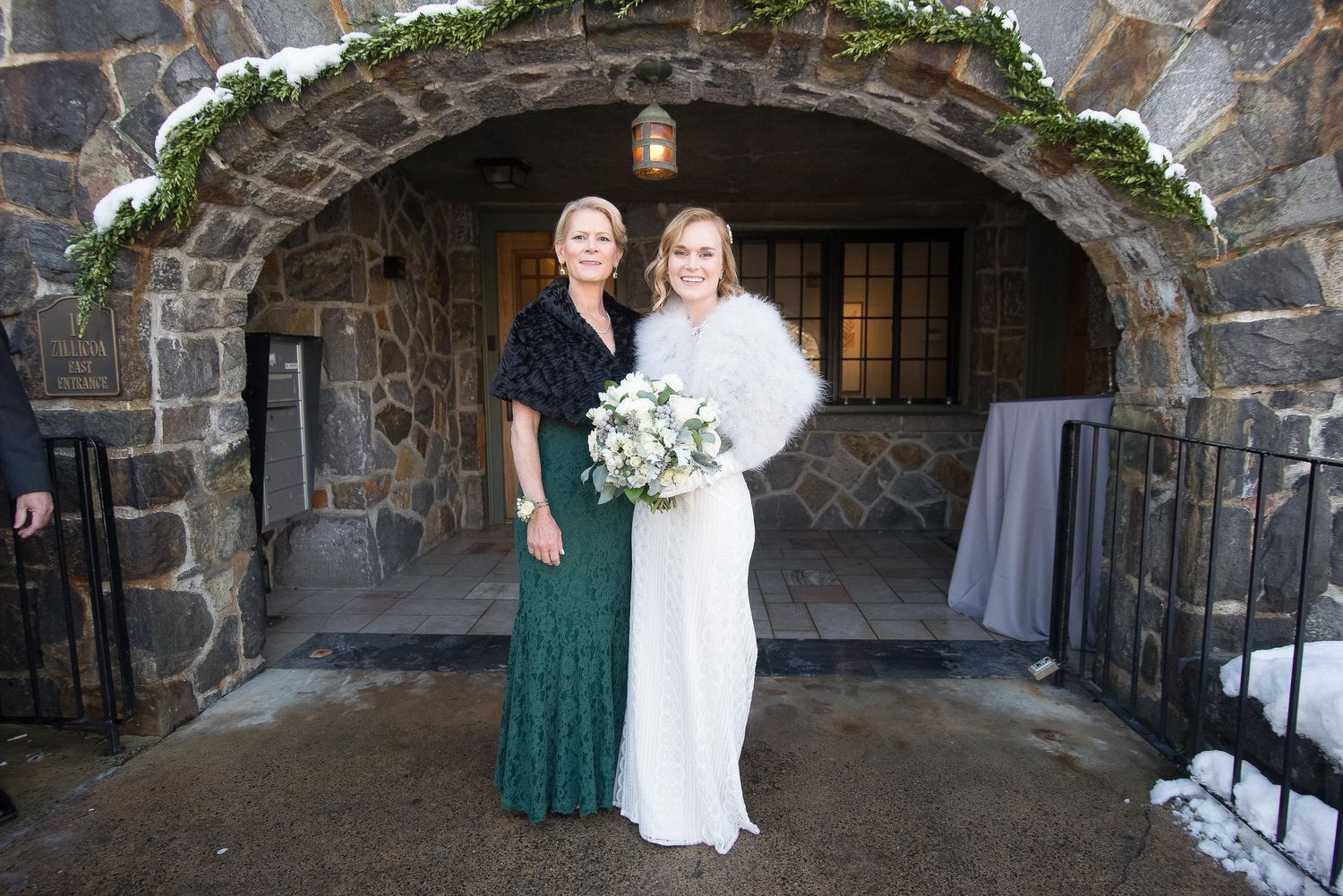 Bride in white gown with fur stole holds bouquet beside woman in green lace dress under stone arch in winter.