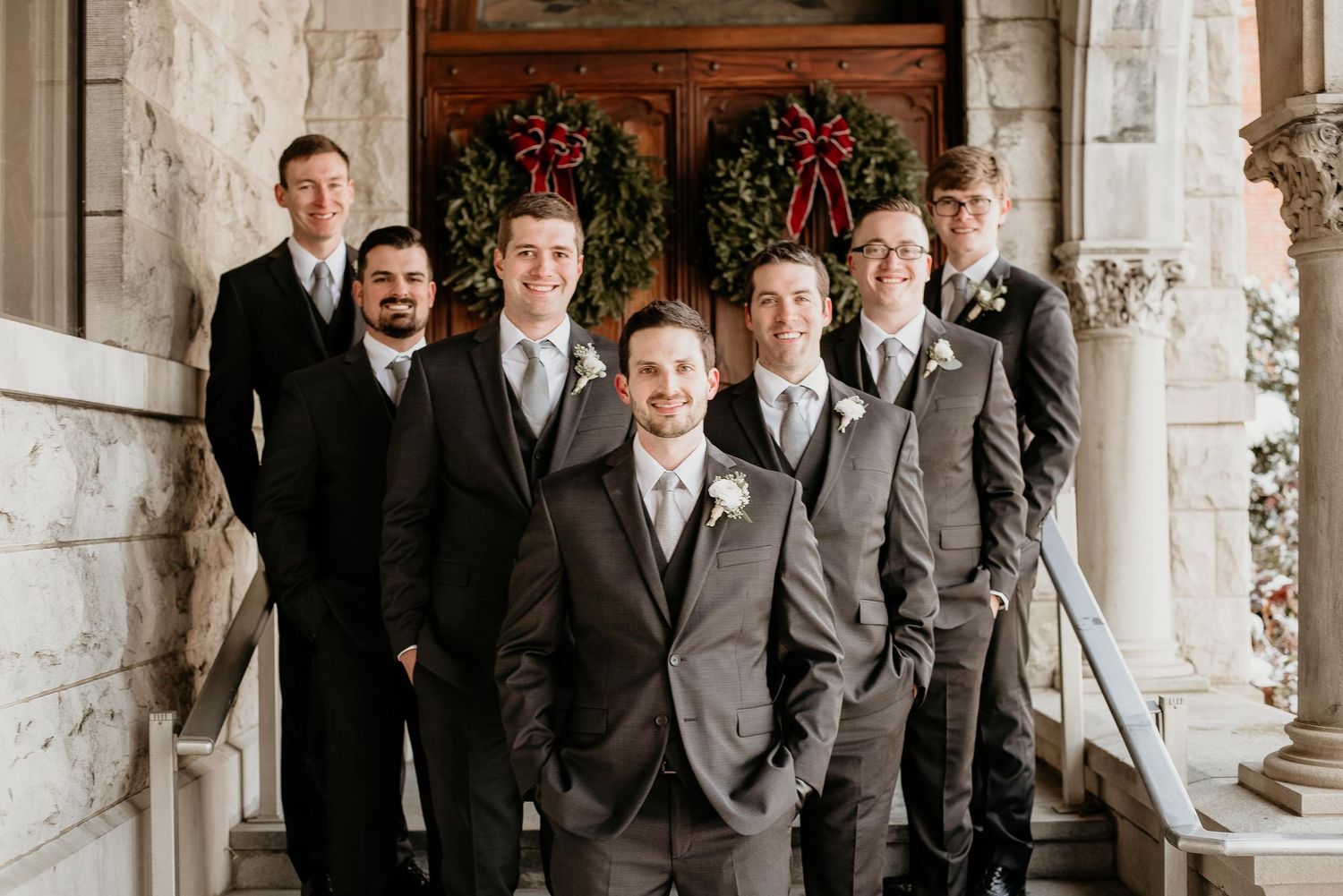 Groom and groomsmen in gray suits pose on stone church steps with Christmas wreaths on wooden doors.