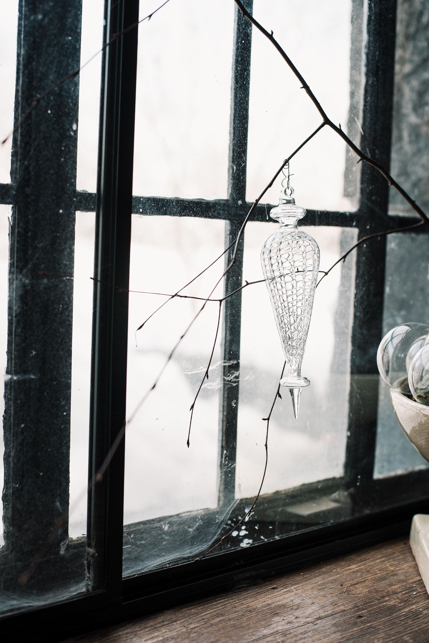 A delicate glass ornament hanging from a bare branch against a rustic multi-pane window with natural light.