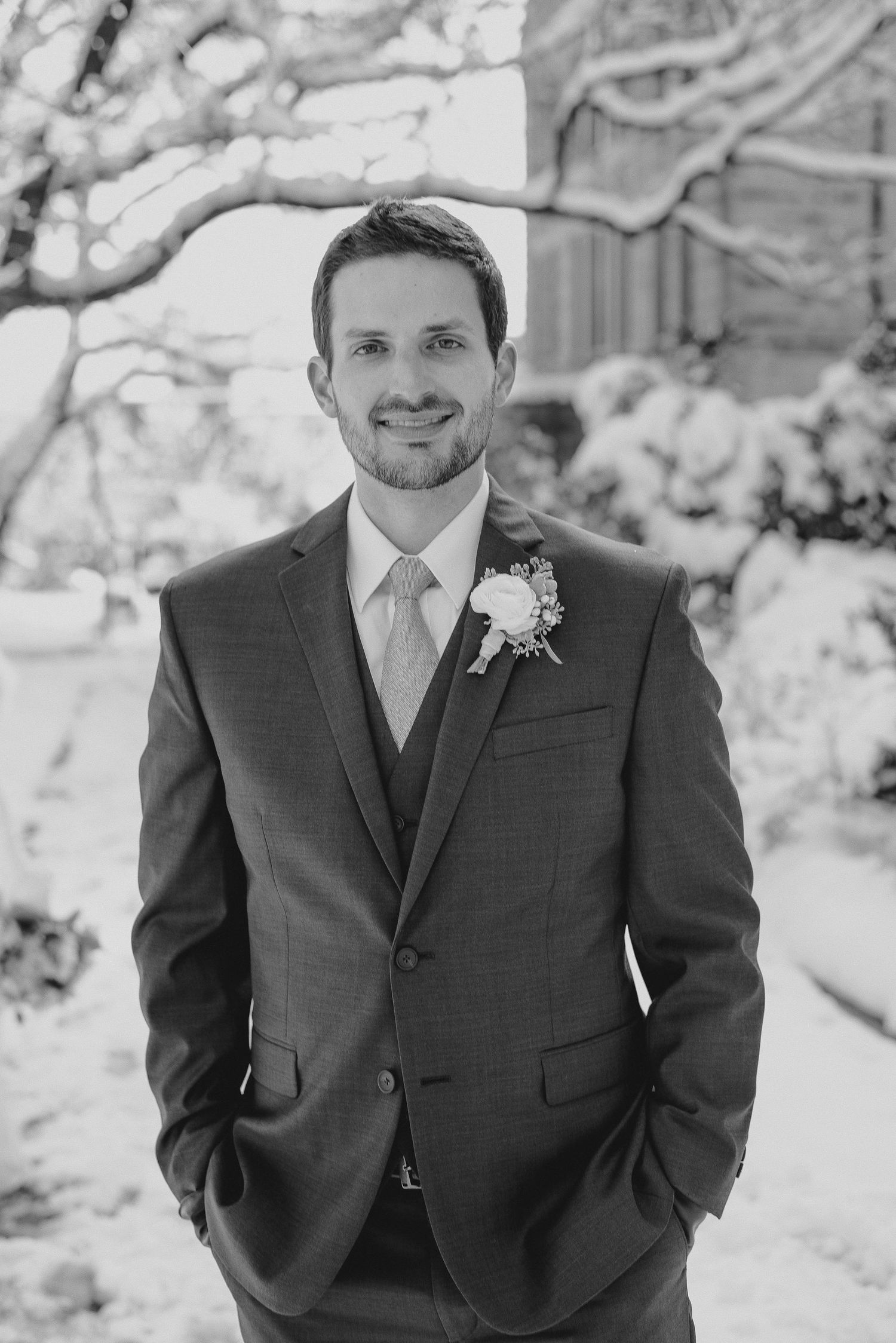 Smiling groom in a three-piece suit with boutonniere poses outdoors in a snowy winter setting for a wedding portrait.