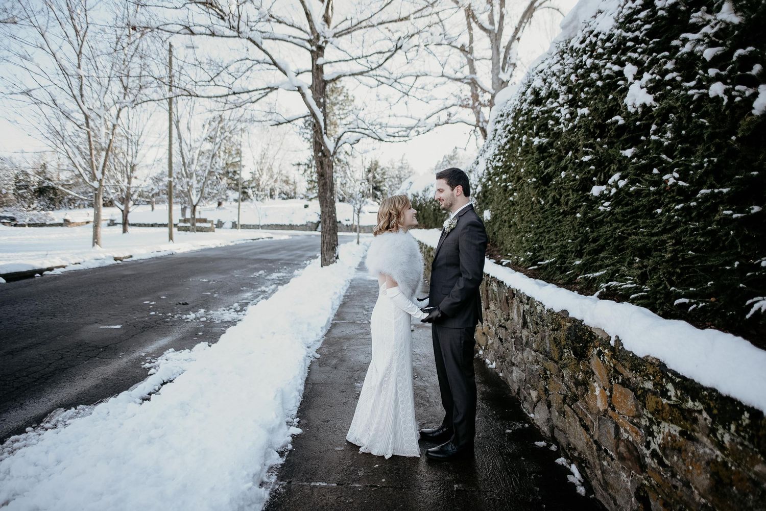 Bride and groom share a romantic winter moment on a snowy sidewalk beside a stone wall.