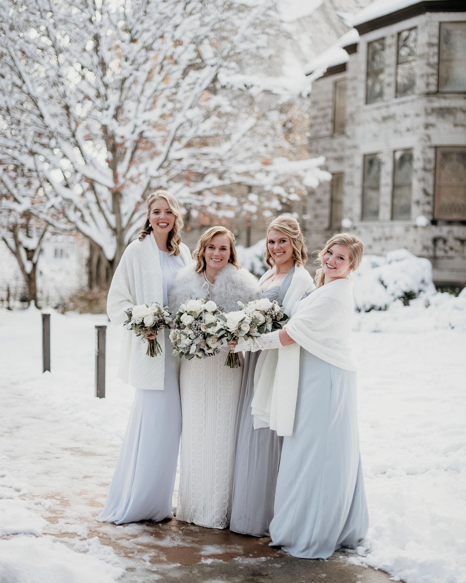 Bride and three bridesmaids in light blue gowns and white wraps holding bouquets outdoors in a snowy winter wedding setting.