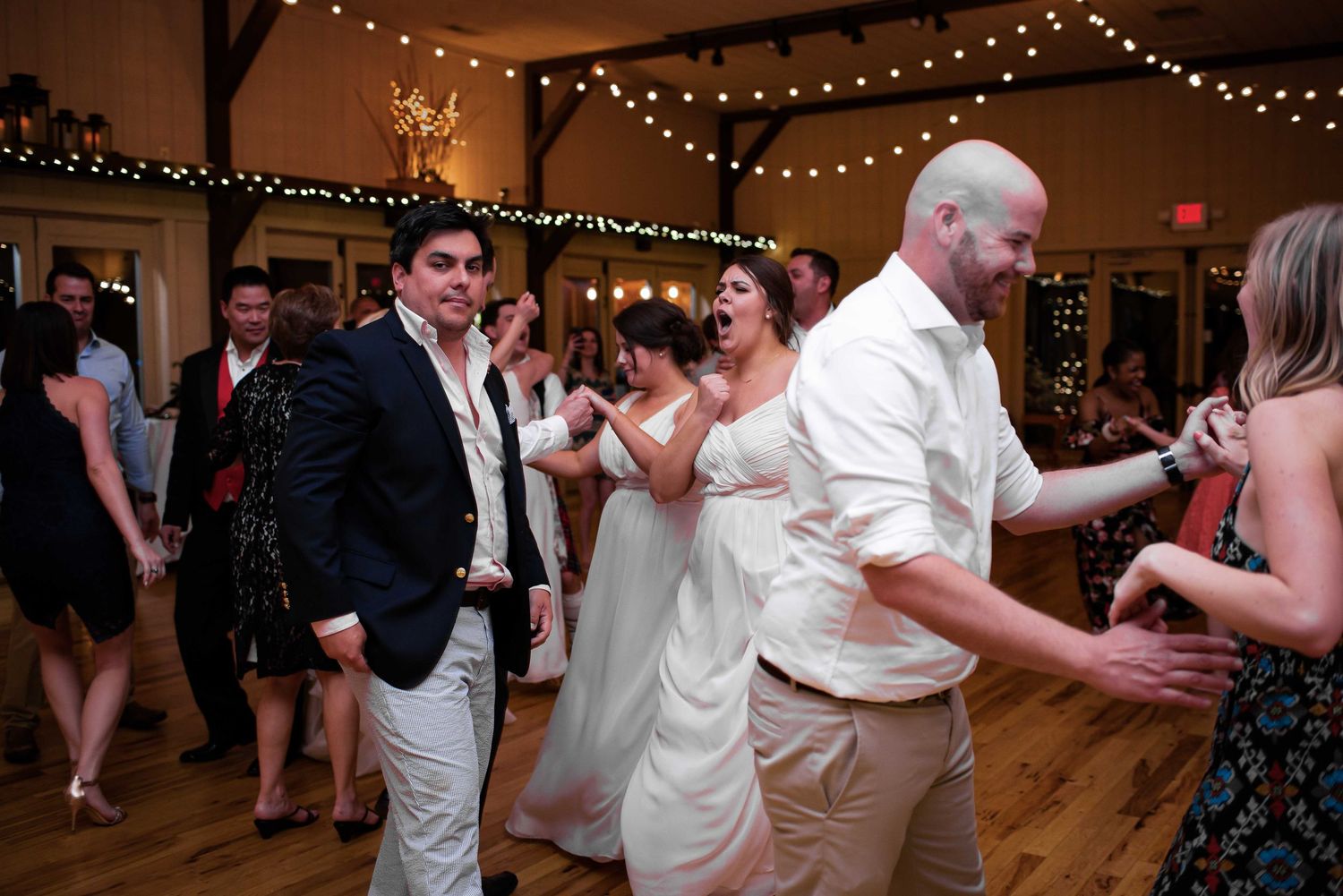 Bride and groom dancing joyfully with guests at a wedding reception with warm string lights.