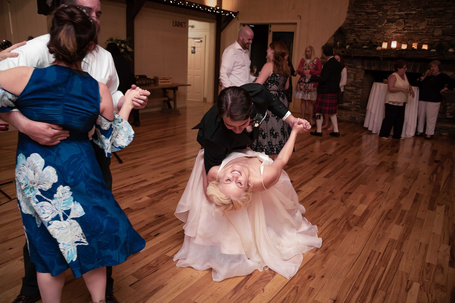 Couple dancing at wedding reception, groom dipping bride in pink dress on wooden dance floor.