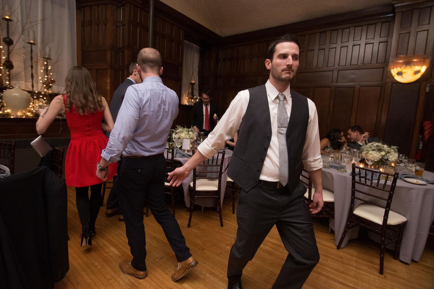 Guests dancing at an elegant wedding reception in a wood-paneled ballroom with string lights and floral centerpieces.